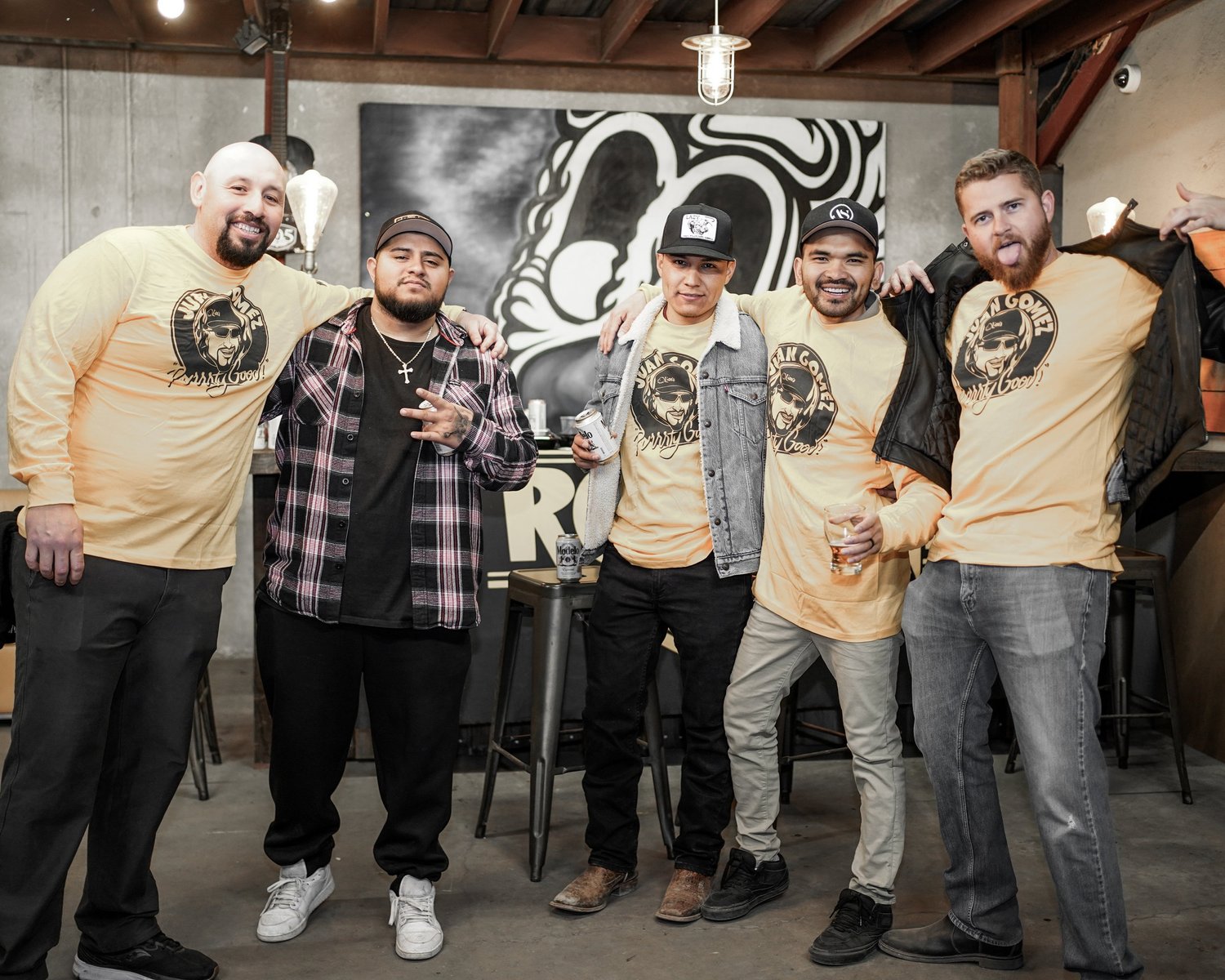 Five men posing together indoors, some wearing matching yellow shirts with a logo, and one in a plaid shirt. They appear to be at a social gathering or party with drinks in hand, smiling and making gestures.