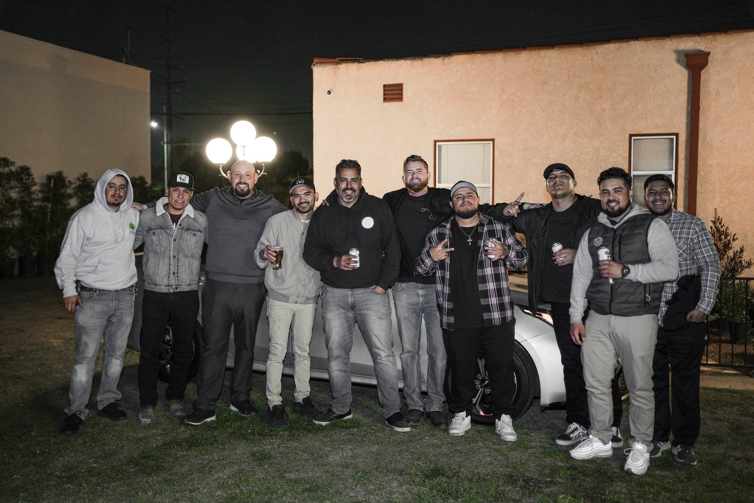 Group of eleven men standing outside at night, posing for a photo with drinks, in front of a building and a white car.