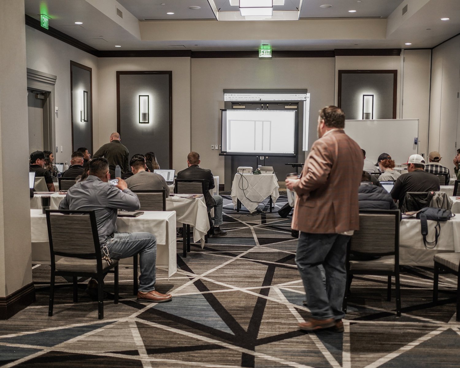 A conference room filled with attendees seated at tables, some working on laptops, facing a large screen at the front of the room. A man in a brown jacket is walking in the foreground.