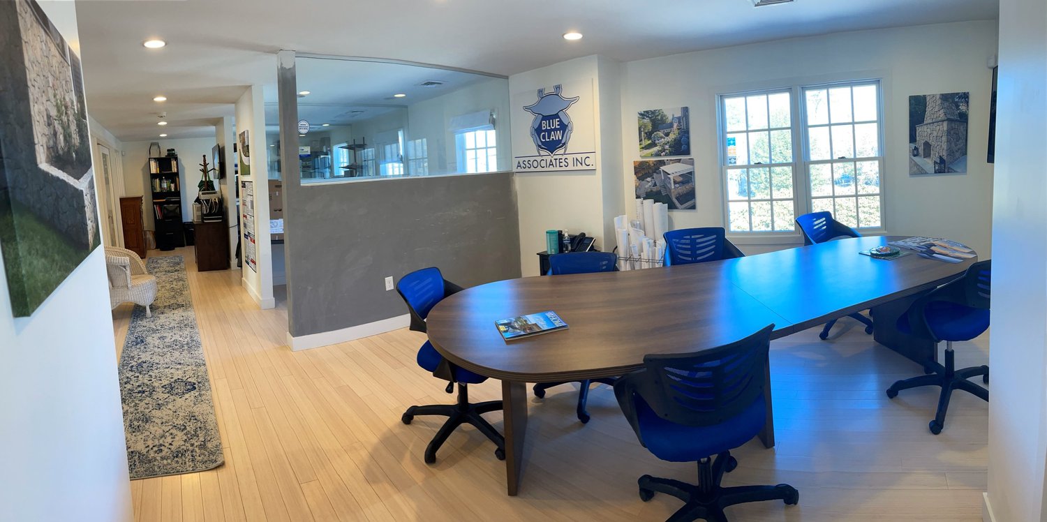 Office conference room with a large wooden table surrounded by blue chairs, windows allowing natural light, and walls decorated with framed pictures and a 'Blue Claw Associates Inc.' sign.