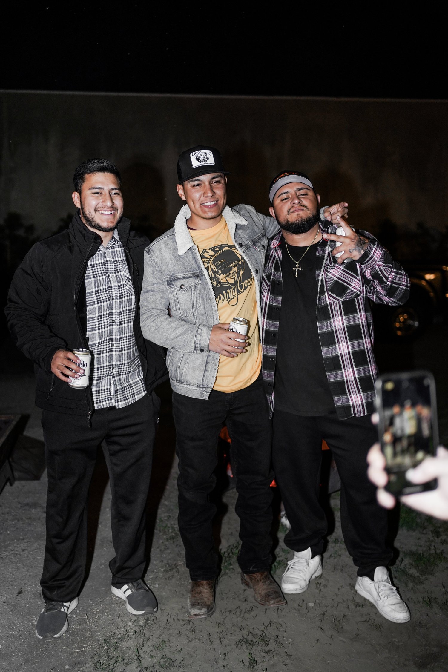Three young men standing at night, smiling, holding drinks, with one making a peace sign, and someone in the foreground taking a photo of them.