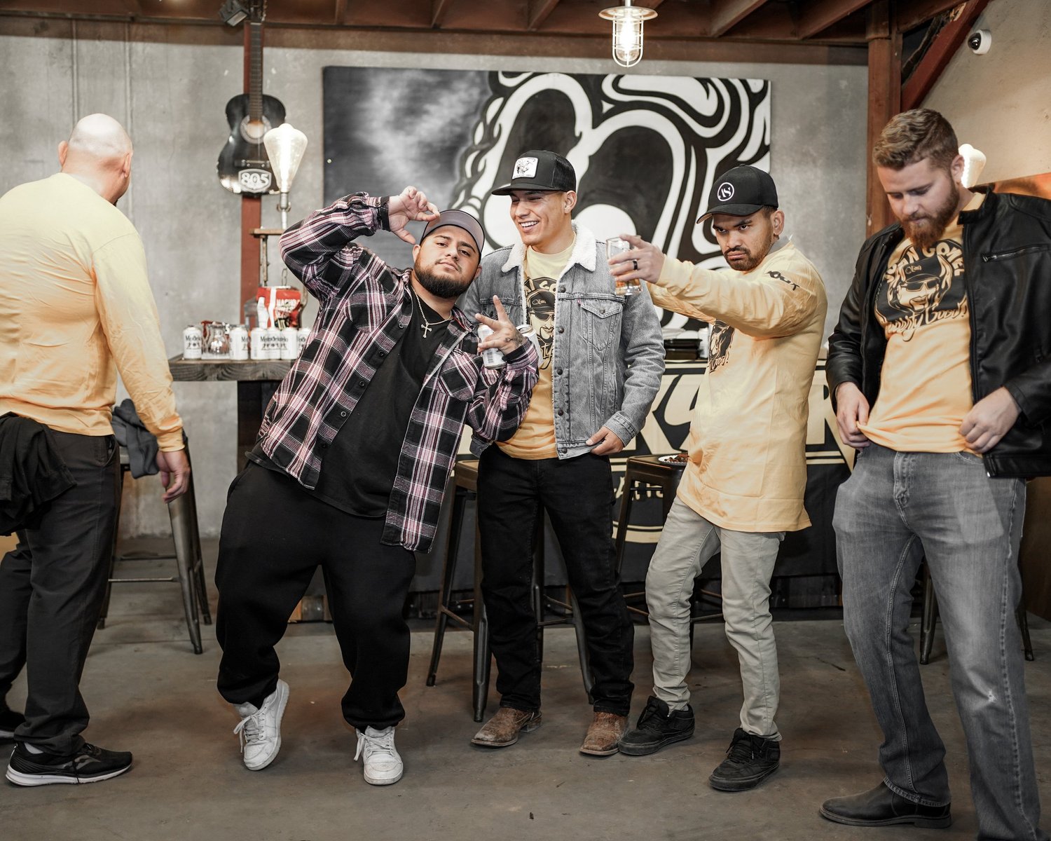 Group of five young men at a bar or club, some making playful poses and holding drinks, in a casual and stylish setting with musical instruments and artwork in the background.