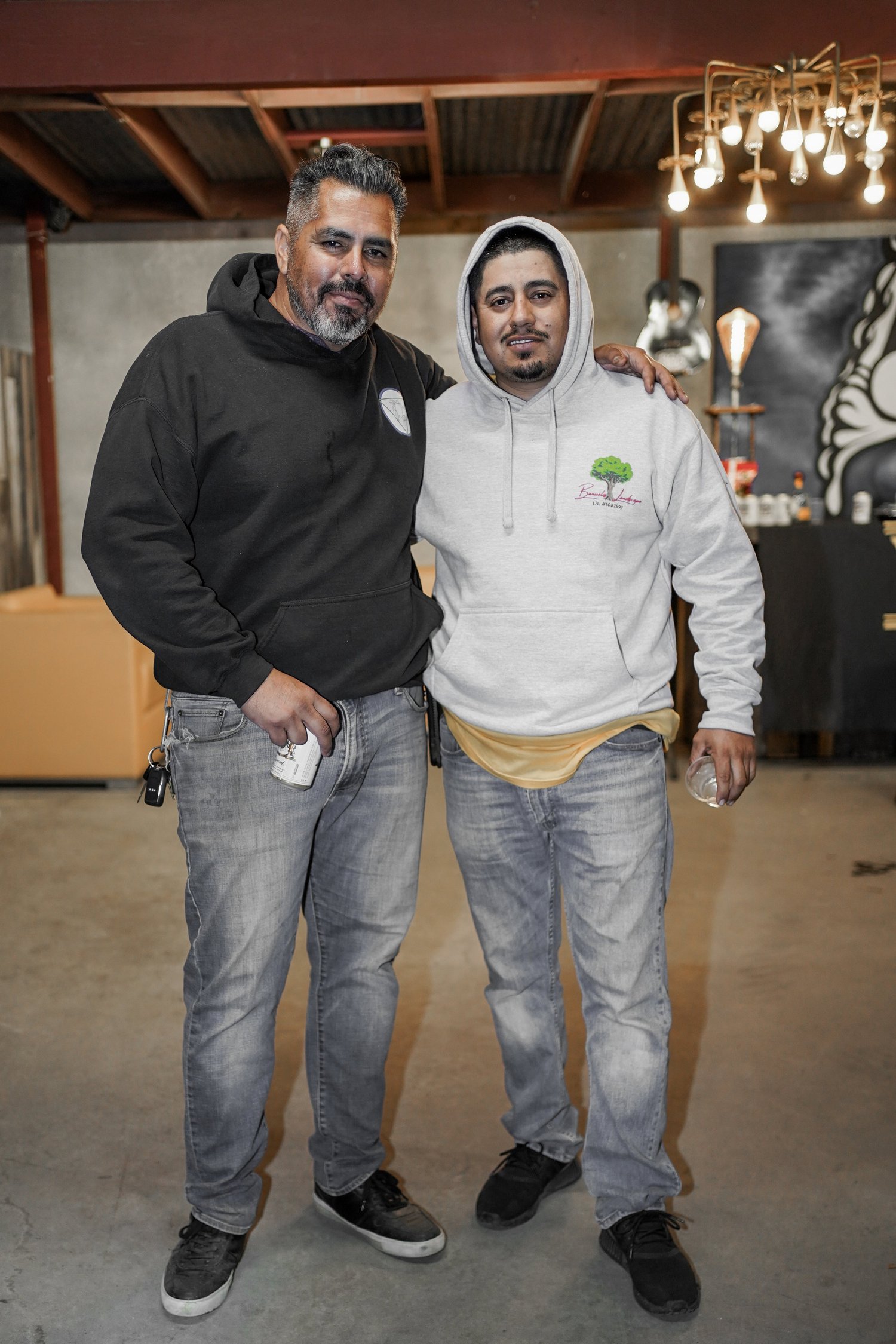 Two men stand together with arms around each other in an indoor setting, one wearing a black hoodie and the other a gray hoodie, both smiling and holding drinks.