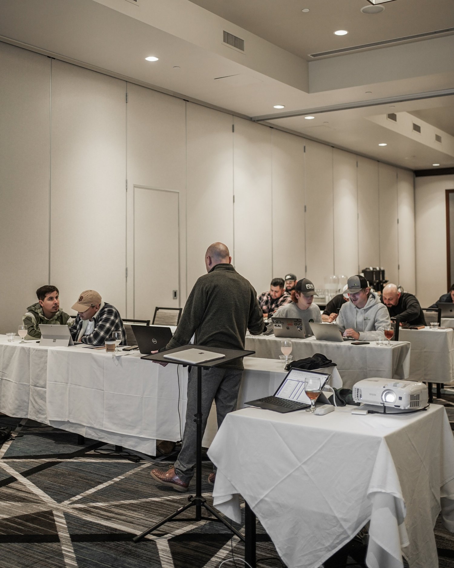 People sitting at tables with laptops in a conference room, with one person standing and speaking.