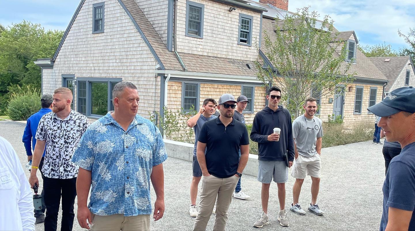 A group of men standing outdoors near a house with a shingle exterior, some wearing casual shirts, shorts, and sunglasses, while one man on the right is wearing a dark cap and navy shirt. Some are holding coffee cups.
