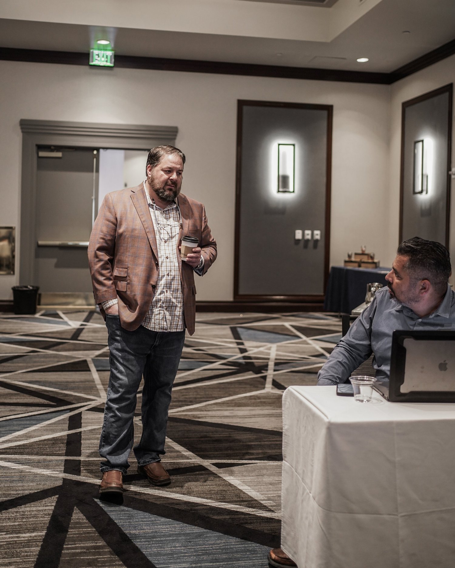Two men are in a conference room, one standing and holding a coffee cup, the other seated at a table with a laptop and glass of water, engaging in conversation.