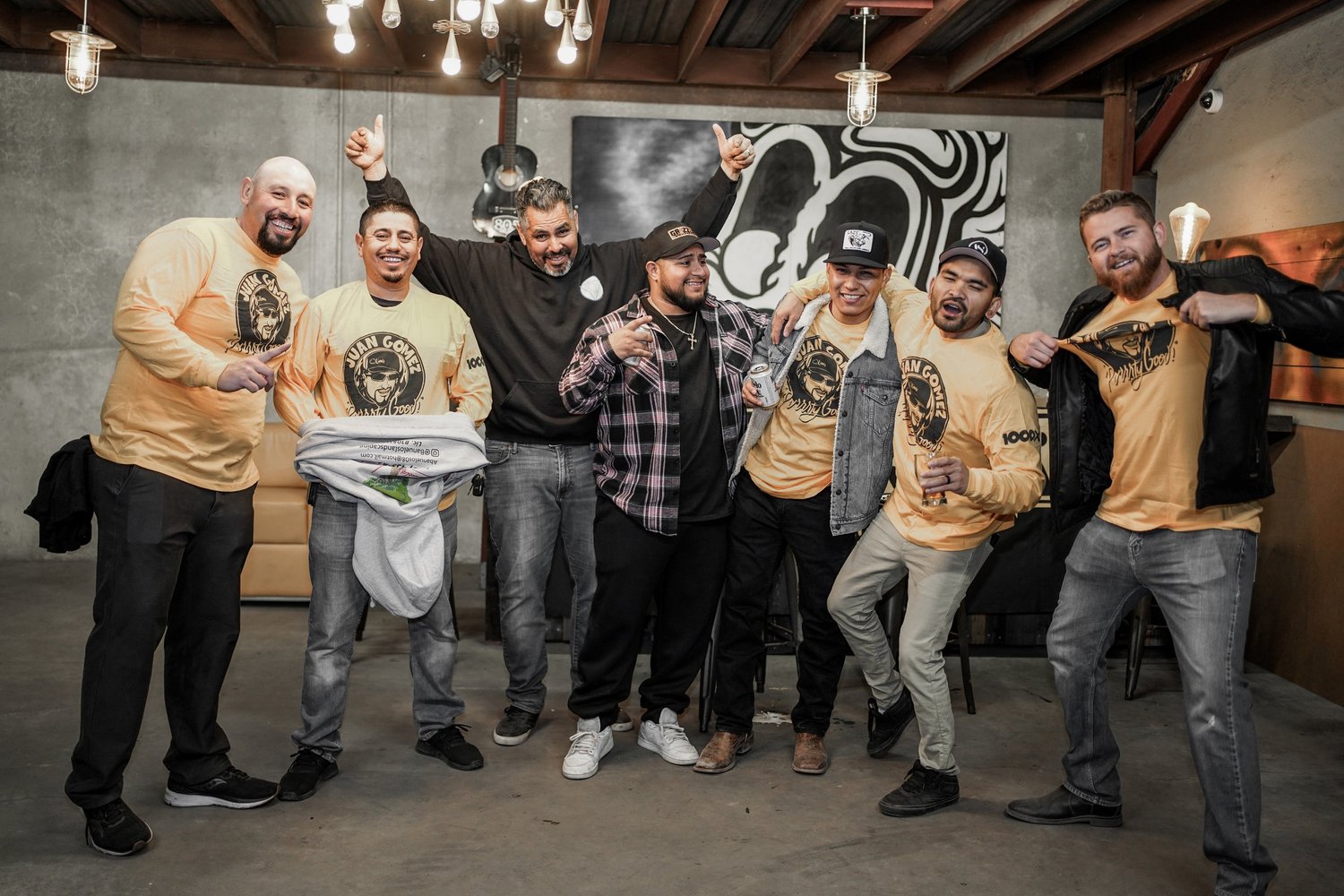 Group of seven men cheering and smiling at a celebration event in a modern loft-style space with gray walls and hanging pendant lights. Some are wearing yellow shirts with a graphic logo, and one man is holding up a shirt, while others are holding dr