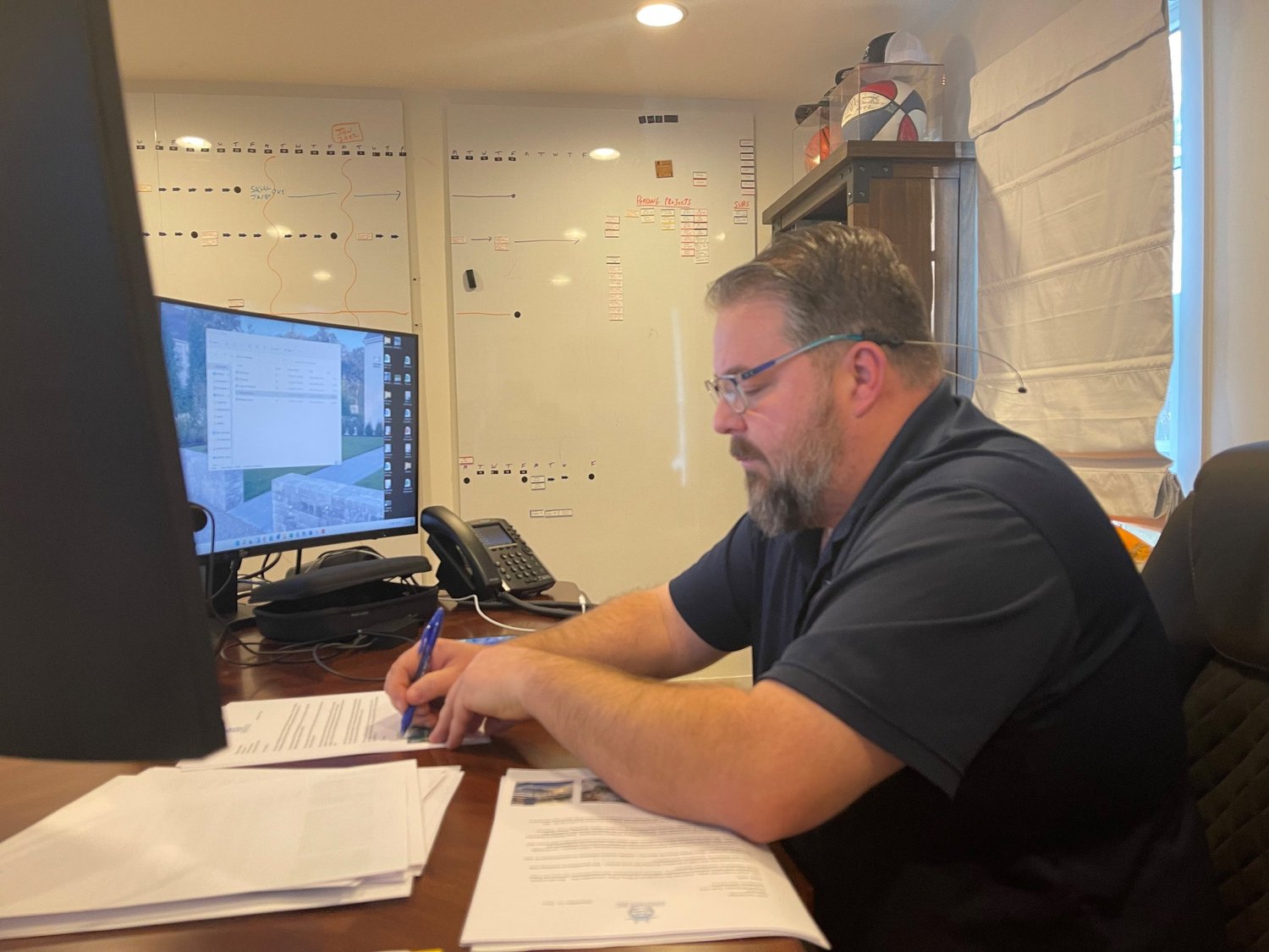 Man with glasses and beard working at a desk with papers, a pen, and a computer in an office, whiteboard with notes and a large window behind him.