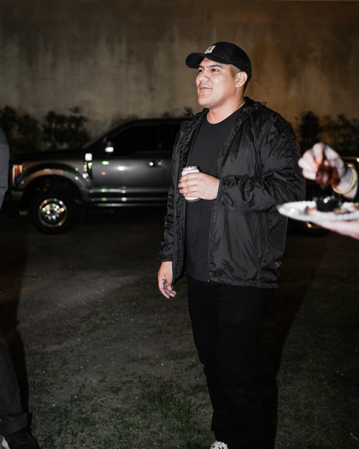 A man wearing a black jacket, black cap, and black pants holding a can of drink, standing outdoors at night with a dark truck in the background.