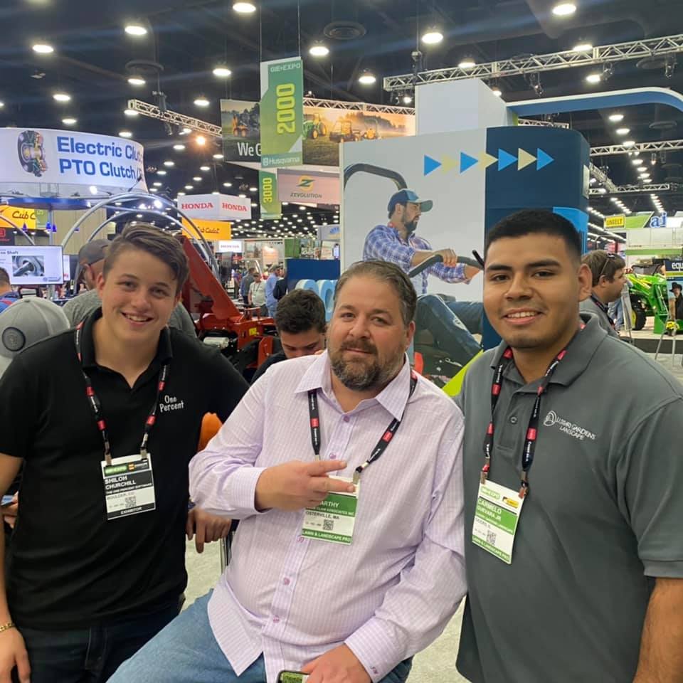 Three men at a convention or trade show, smiling and posing for a photo. The man in the middle points at his chest, with others on either side. They are wearing conference badges around their necks. The background shows booths, banners, and other att