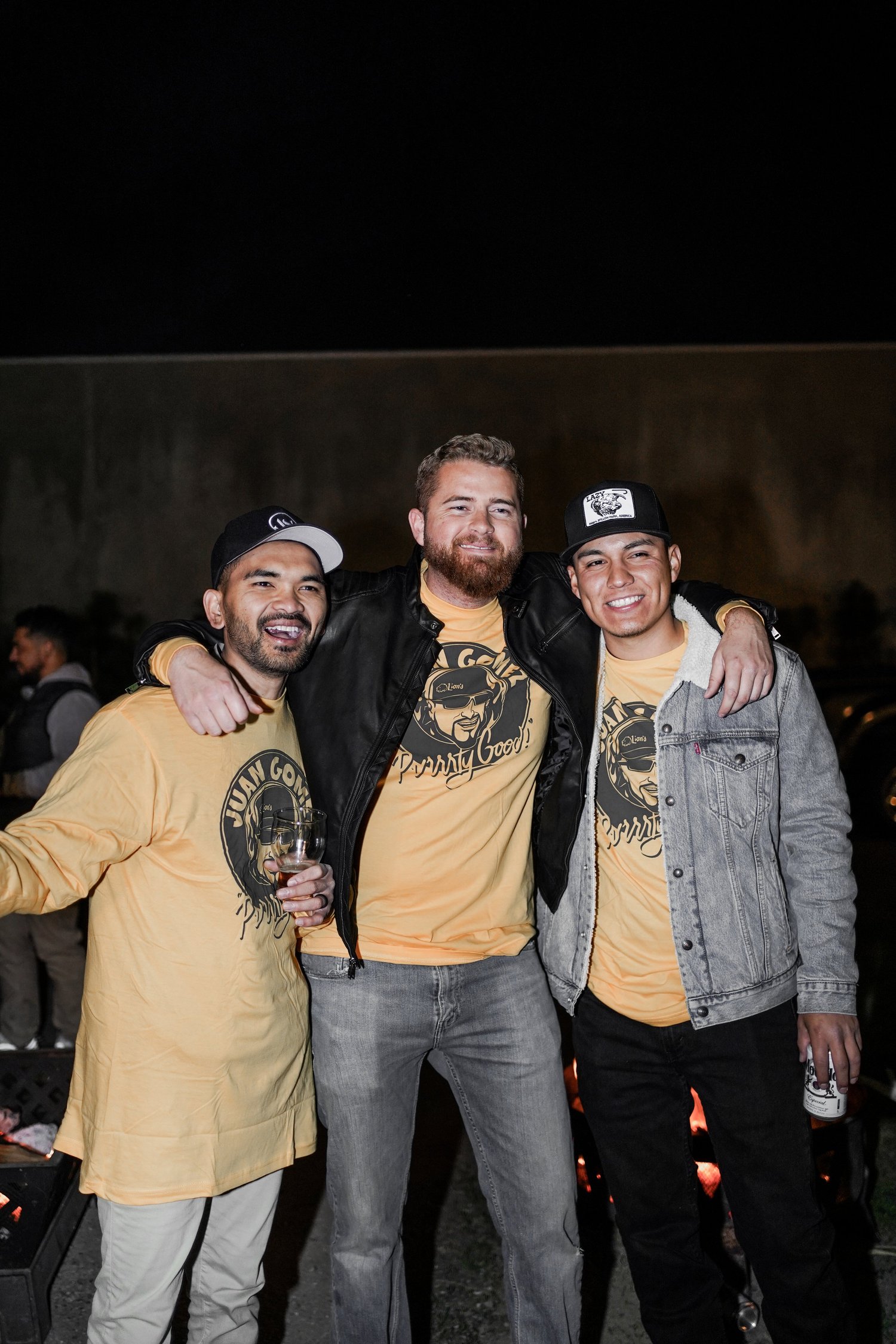 Three men smiling and standing together at night, wearing matching yellow t-shirts with a graphic and text, and casual jackets, with two holding drinks.