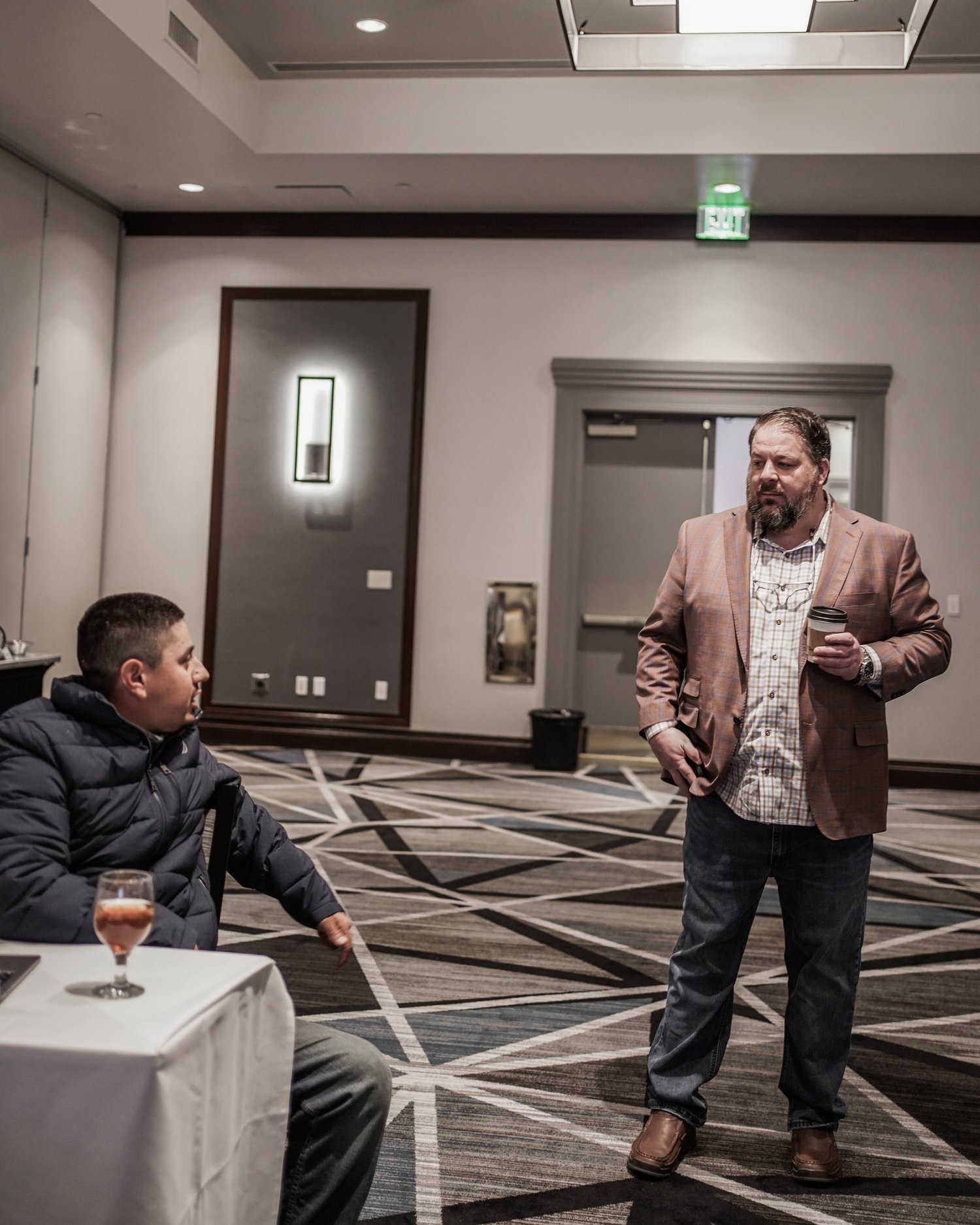 Two men having a conversation in a hotel conference room. One is seated at a table with a drink, the other stands holding a coffee cup.