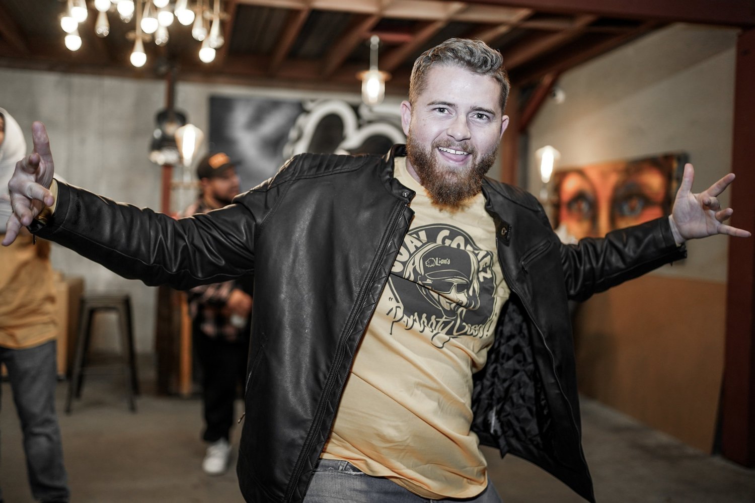 A man with a beard and mustache wearing a black leather jacket and a beige t-shirt with a graphic, smiling and spreading his arms in a casual indoor setting with art on the walls and other people in the background.