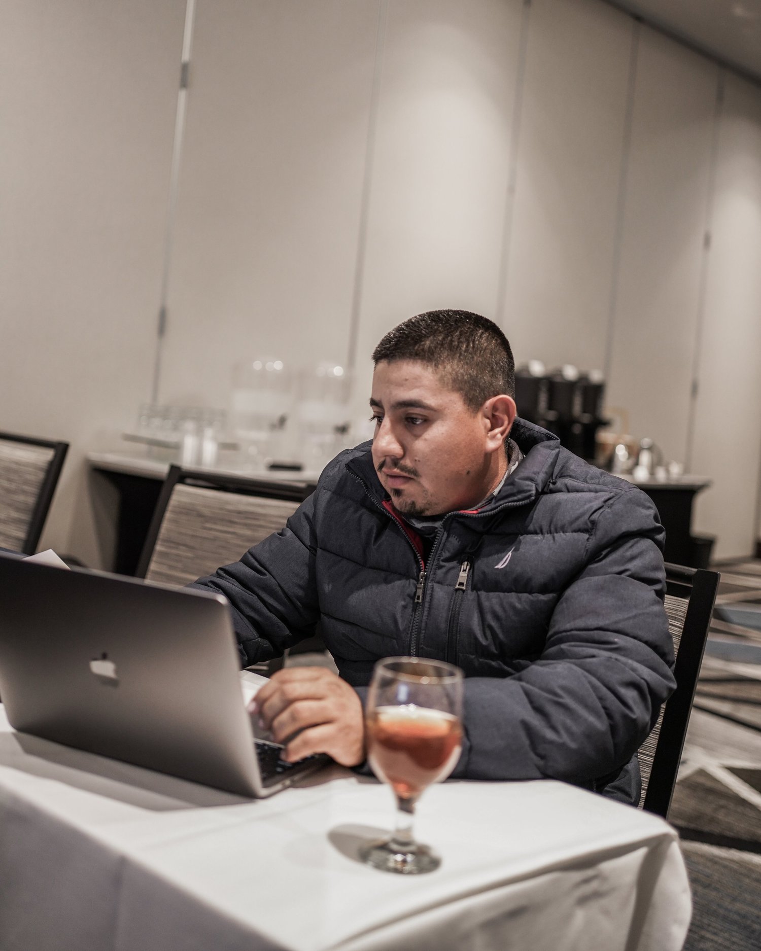 A man in a black jacket working on a laptop at a table in a conference room with a glass of pink drink in front of him.