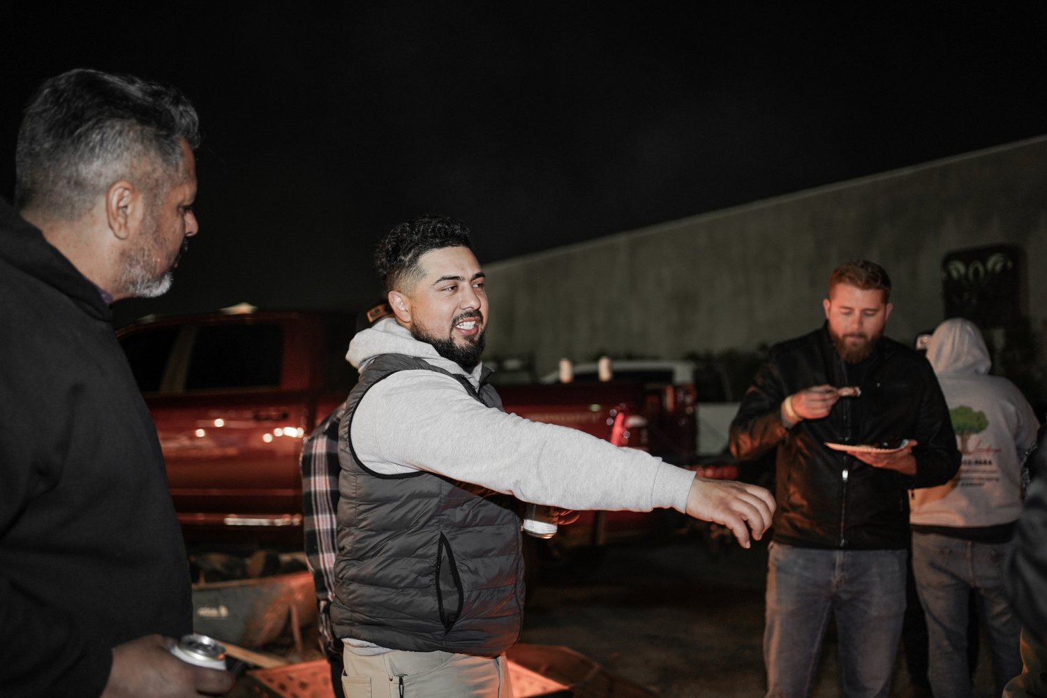 Group of men gathered outdoors at night, one man in a gray hoodie and vest gesturing while speaking, others listen nearby, in a parking lot with trucks in the background.