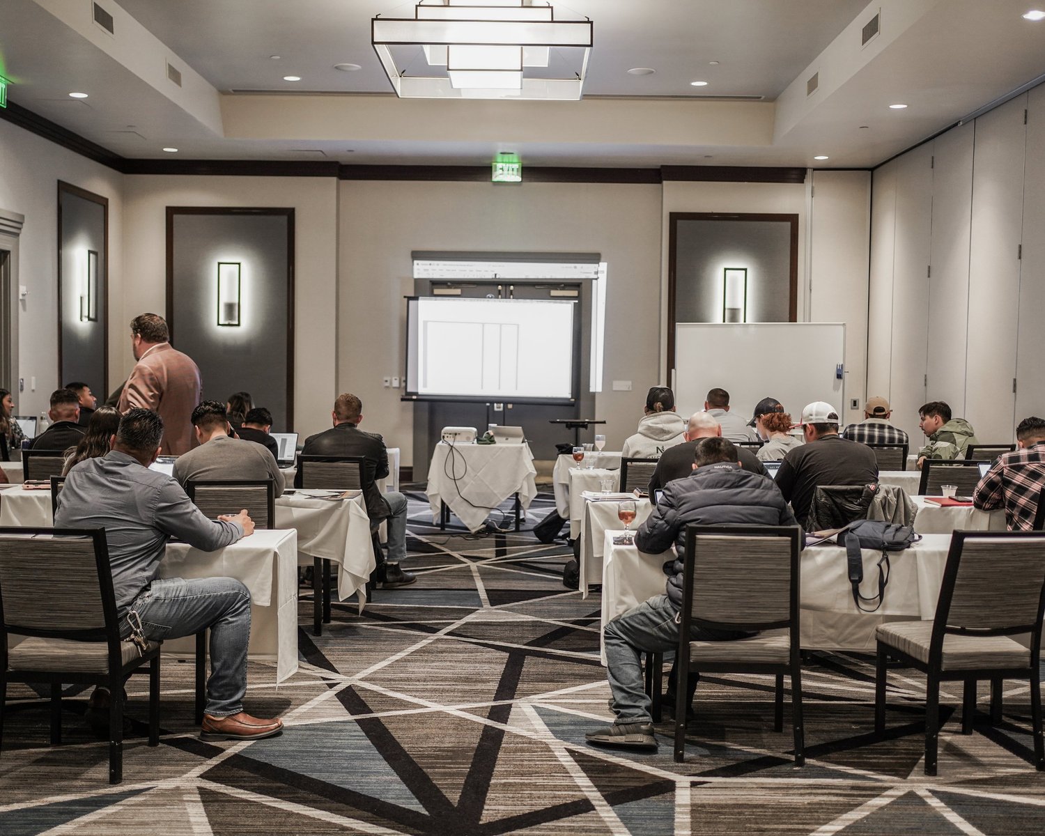 Conference room with attendees seated at tables, a man standing and a projector screen at the front of the room.
