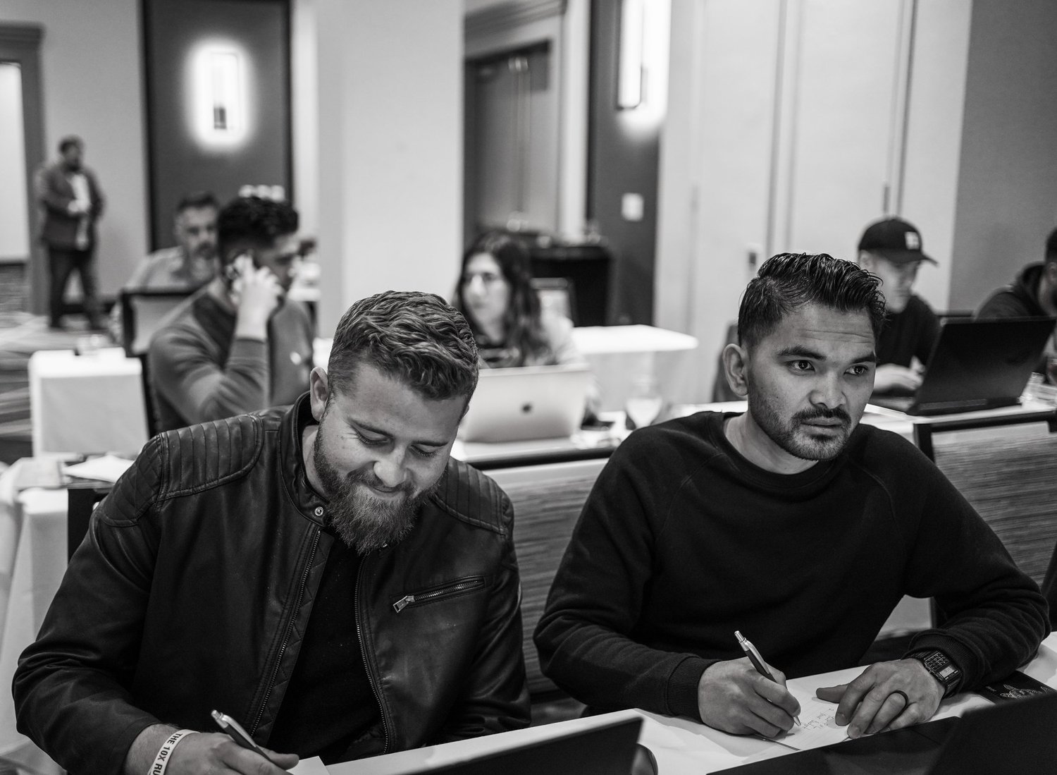 Two men sitting at a table in a conference room, taking notes, with other people working on laptops behind them.