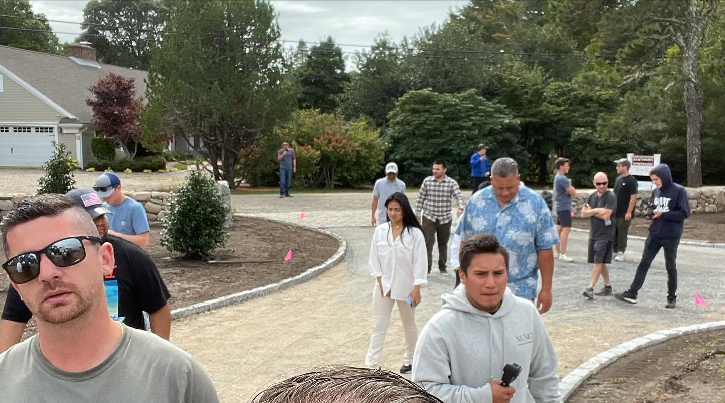 Group of people standing and walking on a gravel path in a neighborhood with trees and houses in the background.