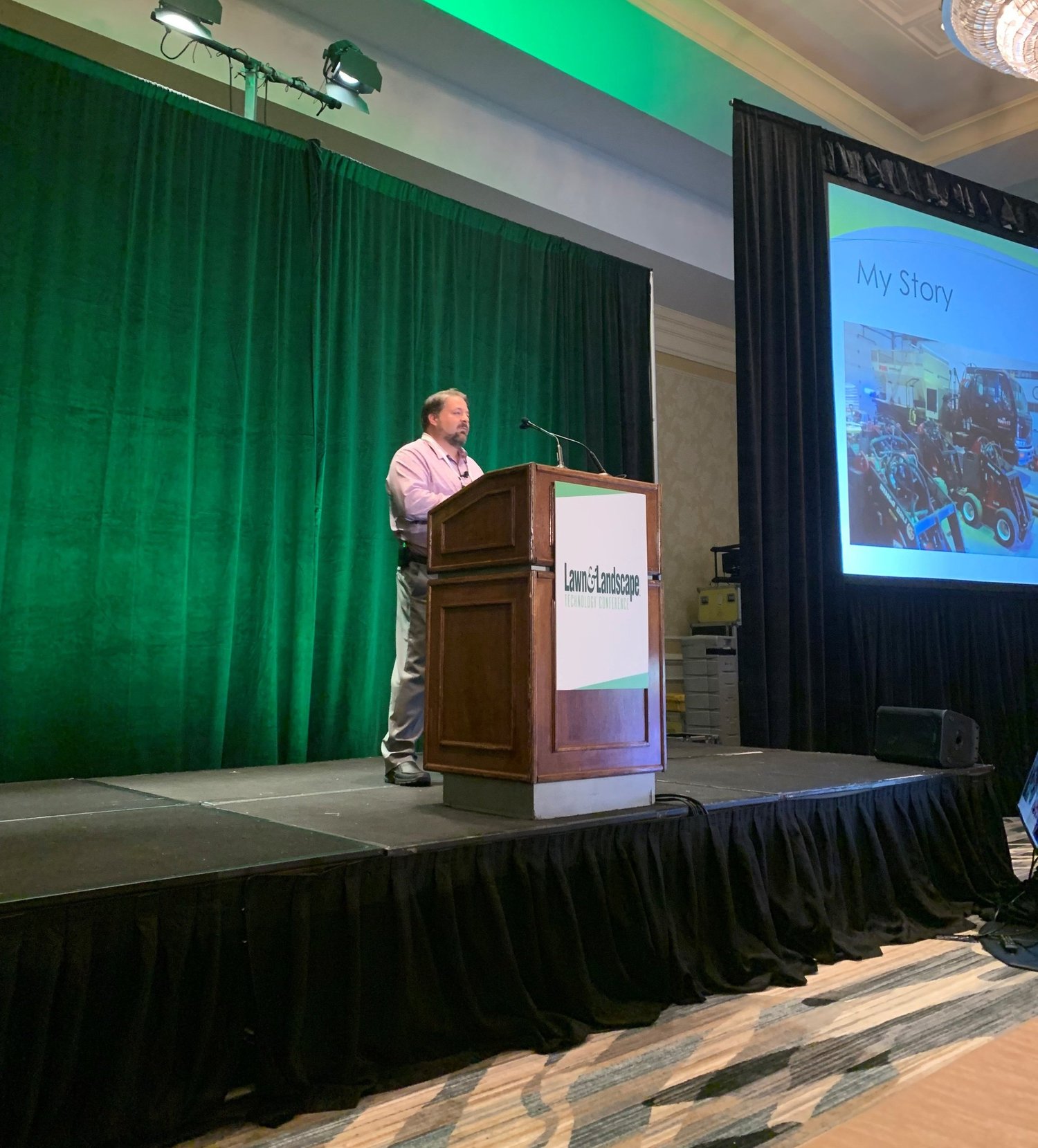 A man giving a presentation at a conference, standing at a wooden podium with a sign that reads 'Lawn & Landscape' and a slide showing a photo of construction equipment and the words 'My Story'.