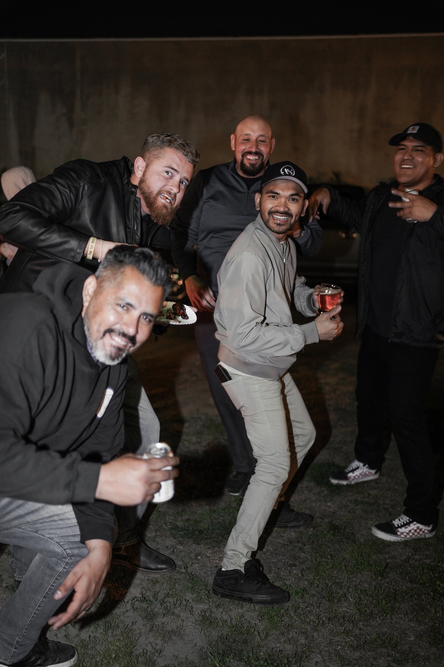Group of five men celebrating with drinks and food at an outdoor gathering at night.
