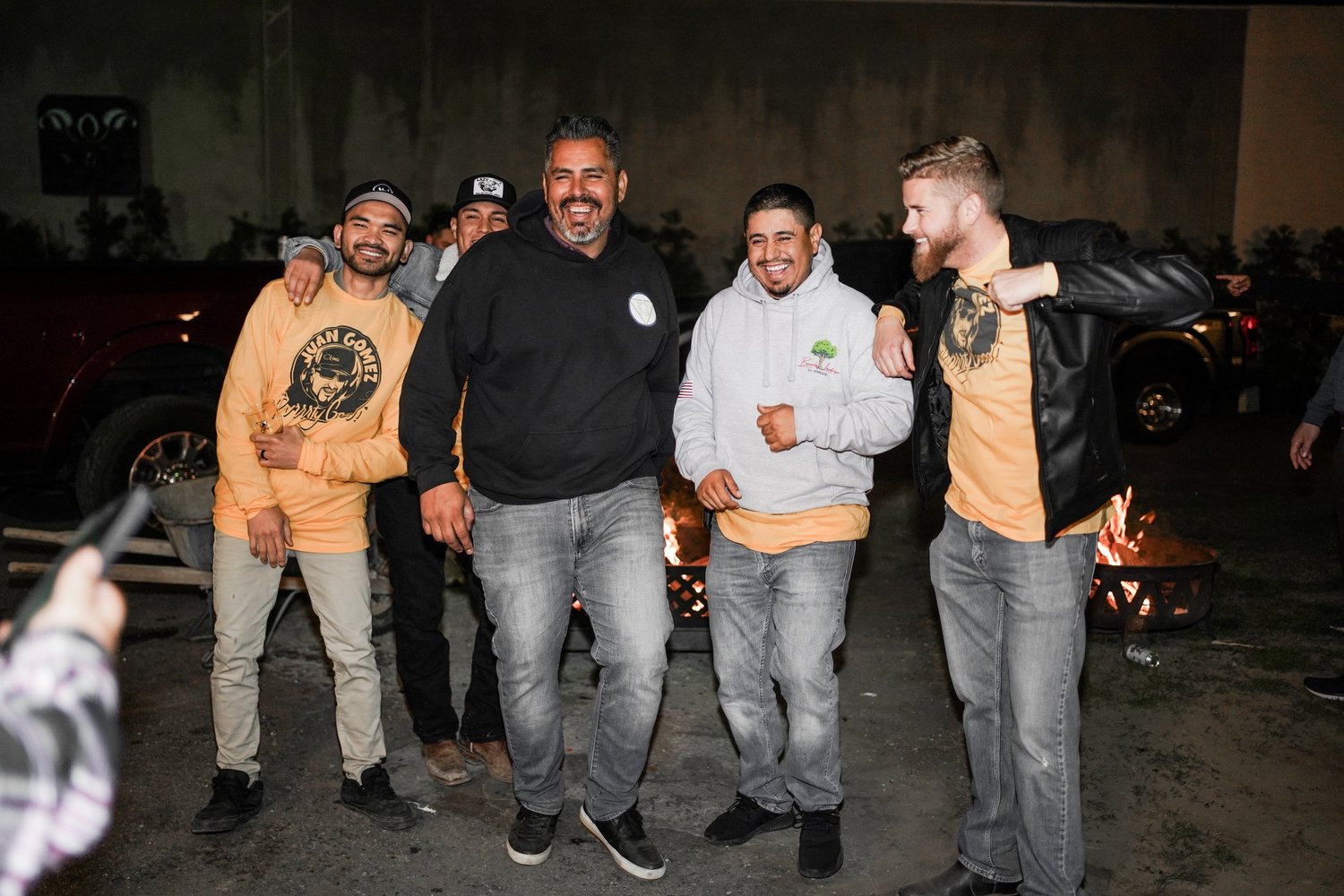 Group of five men standing outdoors near a fire pit at night, smiling and posing for a photo. Three are wearing casual hoodies and jackets, one is wearing a yellow long-sleeve shirt with black graphic, and another in the back is partly visible, also 