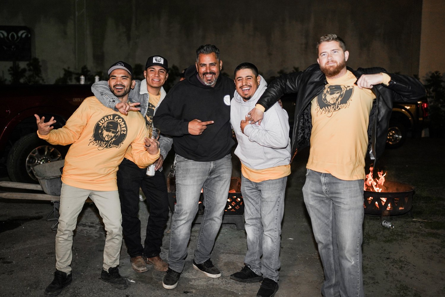 Group of five men smiling and posing in front of a fire pit at night, with trucks and a concrete wall in the background. Some are wearing matching yellow T-shirts with a face and text, while one is wearing a black hoodie and another a black leather j