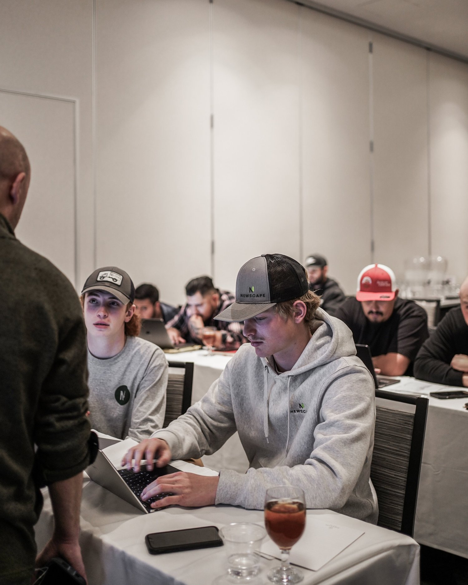 A group of young men in casual clothing and caps sitting at a long table in a conference or meeting room, some looking at their laptops and others listening to a speaker standing in front of them.