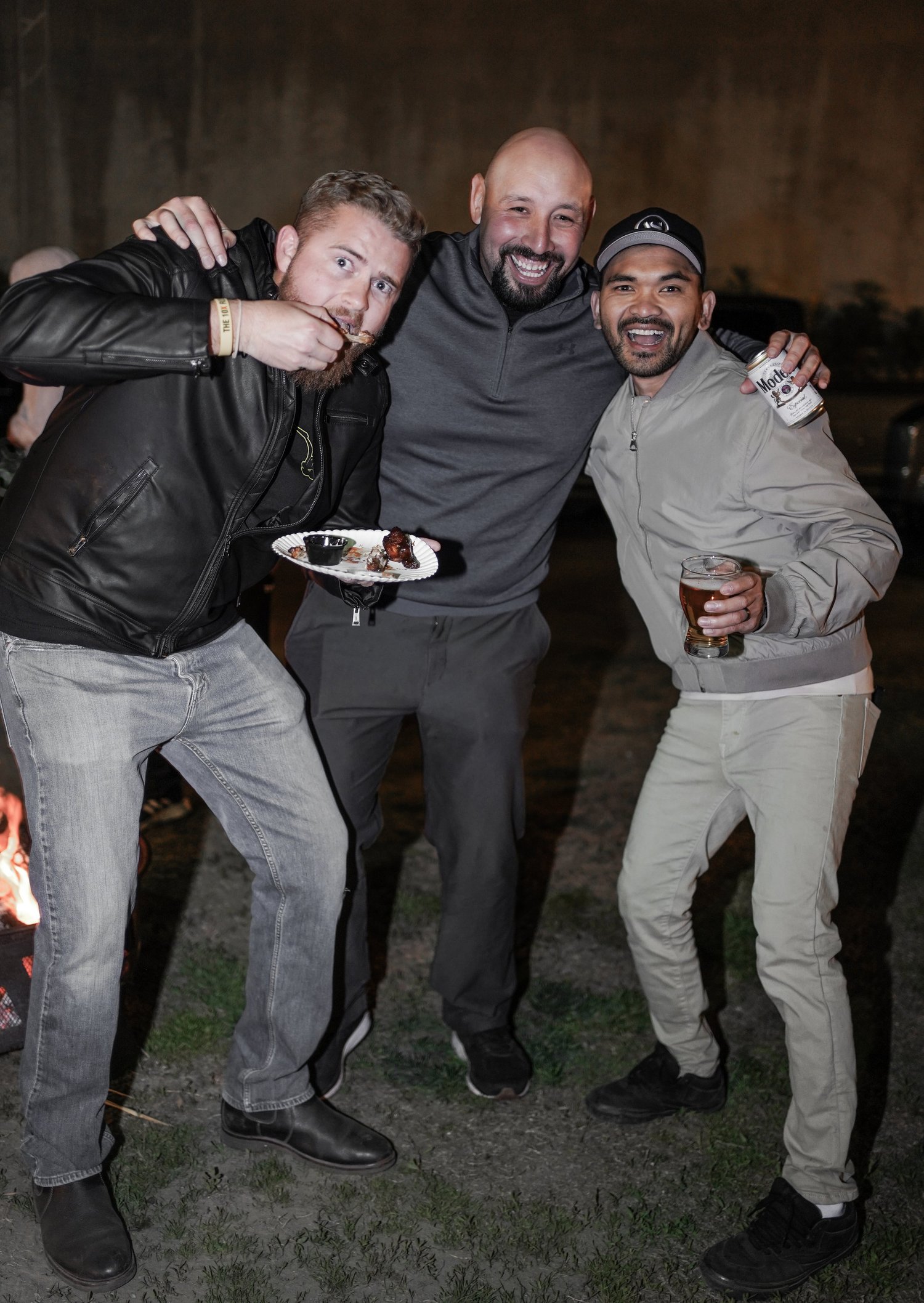 Three men smiling and enjoying a night out. One man is holding a plate of meat and eating, another has a glass of beer, and the third is holding a can of Modelo beer with a glass in front of him. They stand on grass near a fire pit at night.