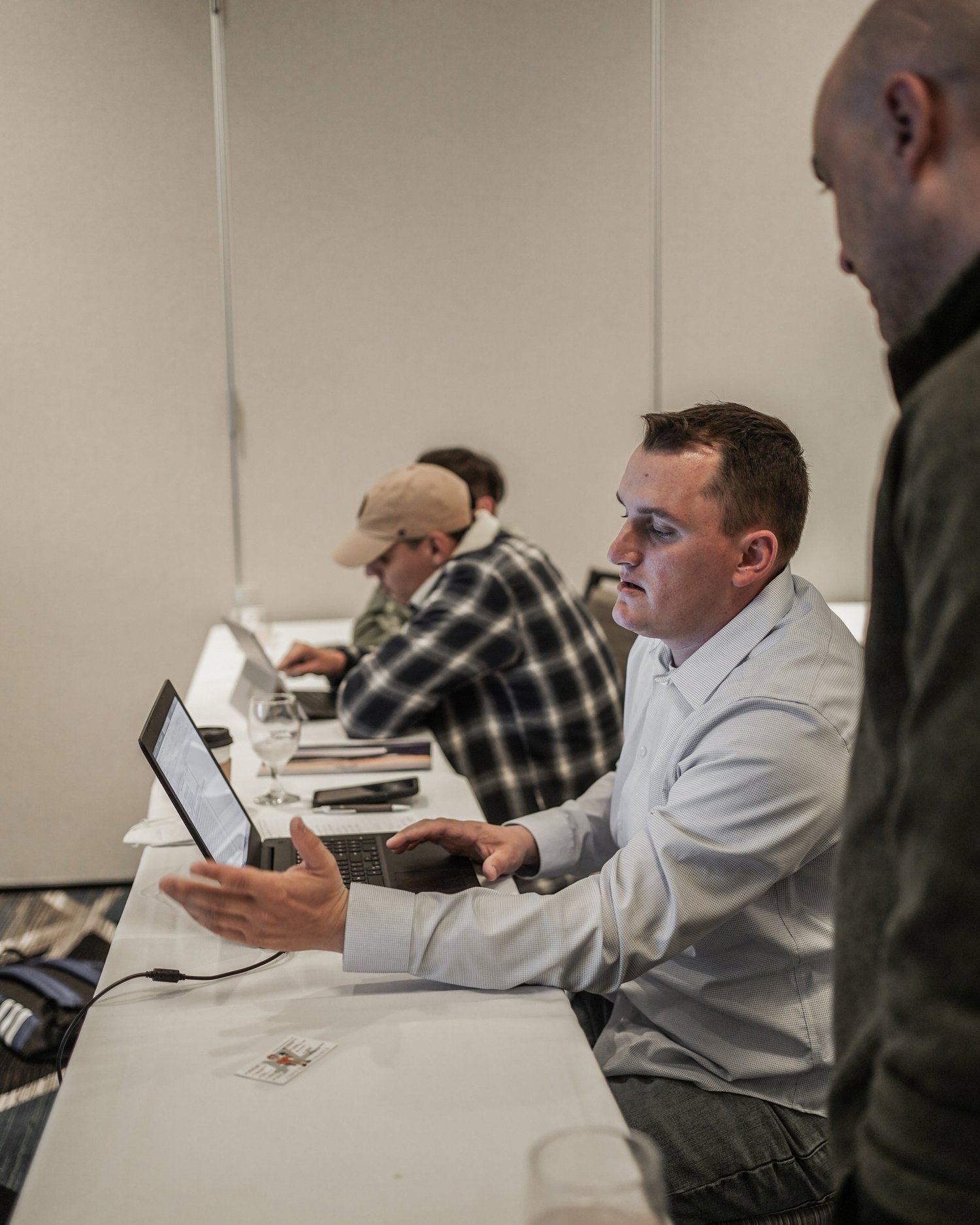 A man in a white shirt demonstrating something on a laptop to another person in a dark shirt at a conference table with several other people working.