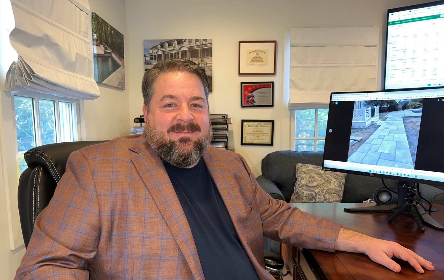 A man with a beard and brown hair, wearing a plaid blazer and black shirt, sitting at a desk in an office. There are windows with white blinds, framed certificates and photos on the wall, a computer monitor displaying a patio, and a cozy chair with a