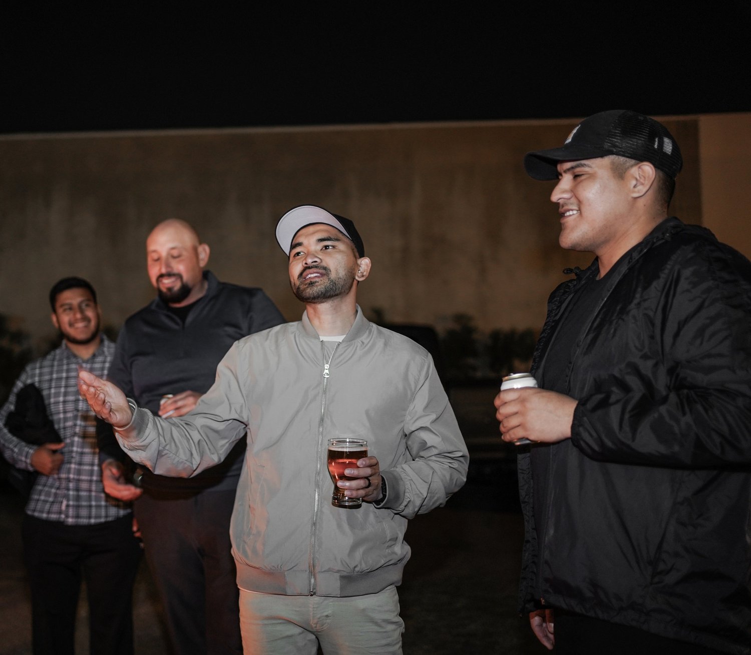 Group of five men socializing outdoors at night, two holding drinks, one gesturing as he talks, smiling and listening.