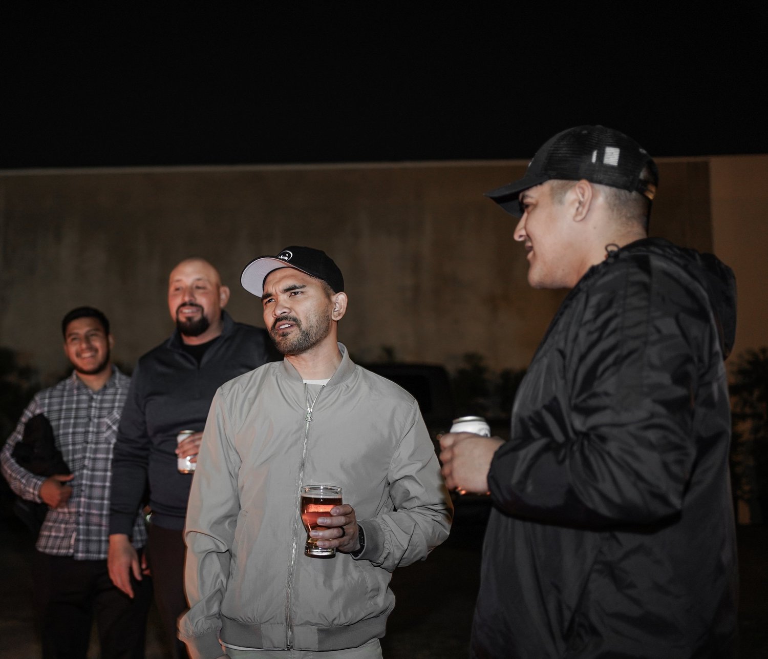 A group of five men at night, socializing outdoors, holding drinks, with a modern building in the background.