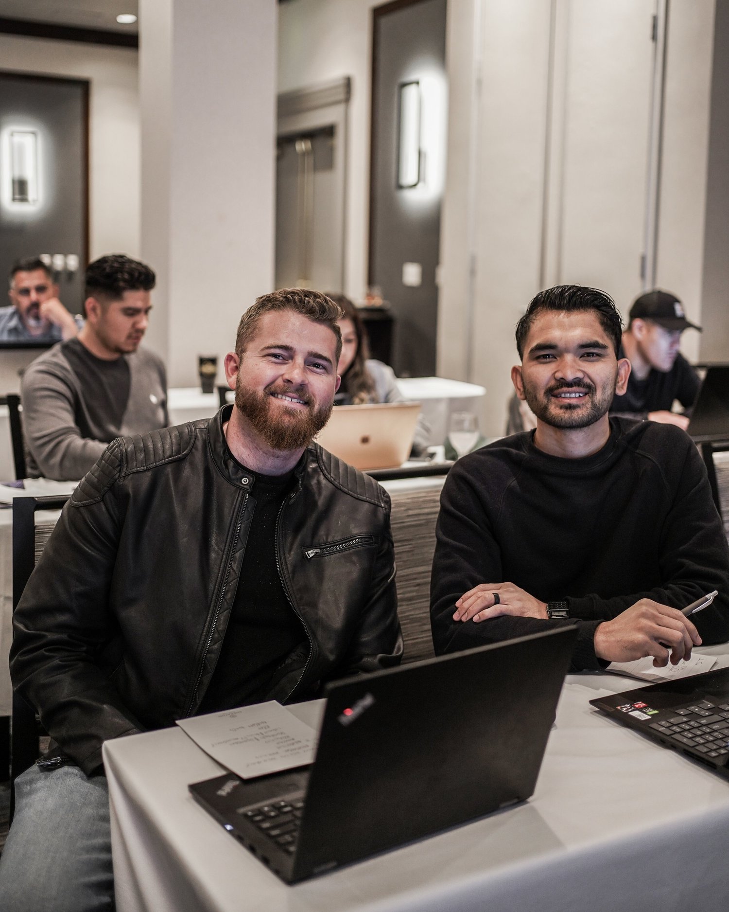 Two men smiling at a conference table with laptops and notes, in a meeting room with other attendees working in the background.