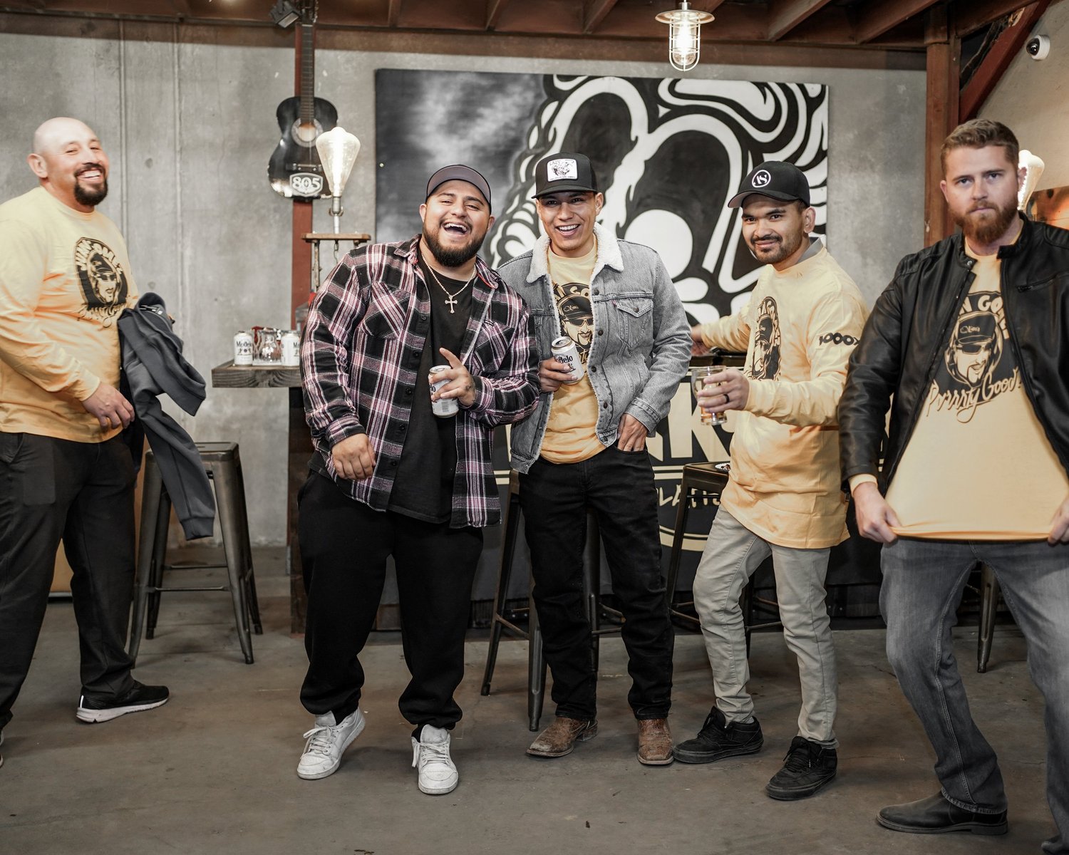 A group of six men smiling and holding drinks in a cozy, industrial-style bar or café with concrete walls, wooden ceiling beams, and modern pendant lighting. Some men are wearing matching yellow T-shirts with a graphic design.