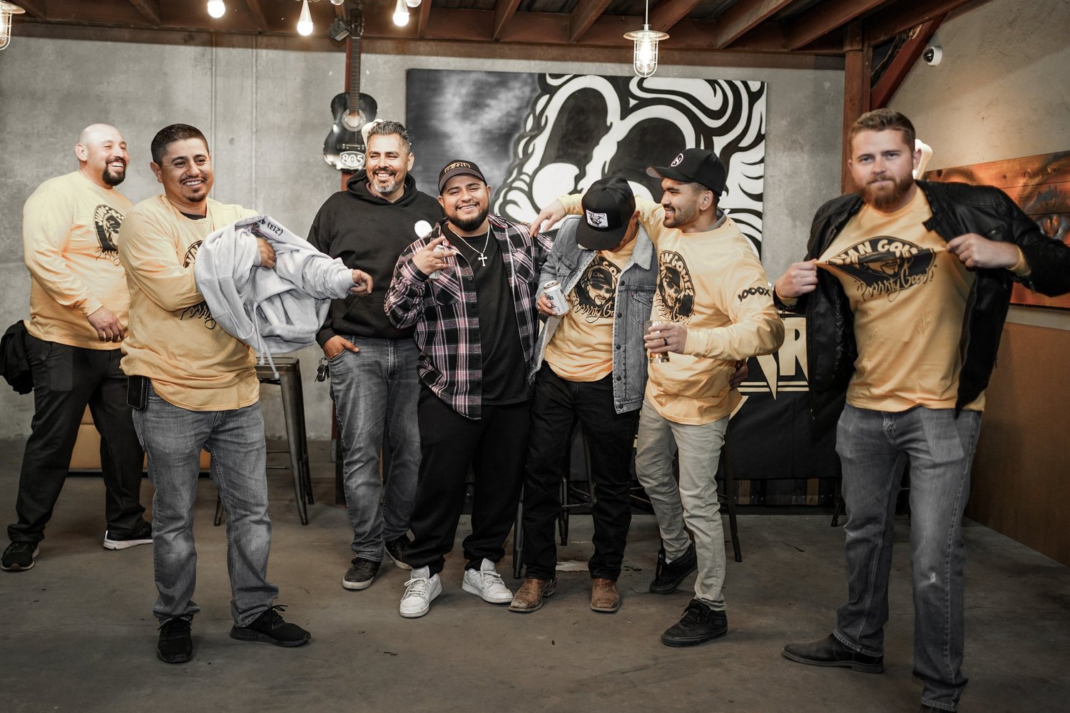 A group of eight men is standing together indoors, smiling and posing for the photo. They are wearing casual clothing, with some in matching tan shirts with a black and gold logo, and others in various casual outfits. The background features a large 