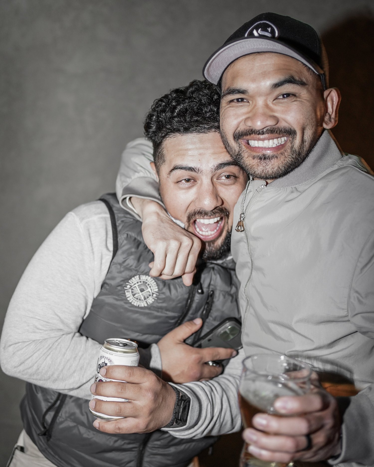 Two men smiling and celebrating together, one holding a drink and the other a can of soda, with a gray background.