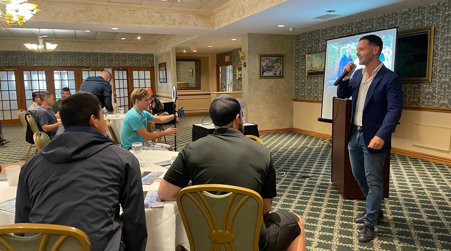 Man in a blue blazer and jeans speaking into a microphone at a presentation in a conference room. Audience members seated at tables, some taking photos or videos, watching him.