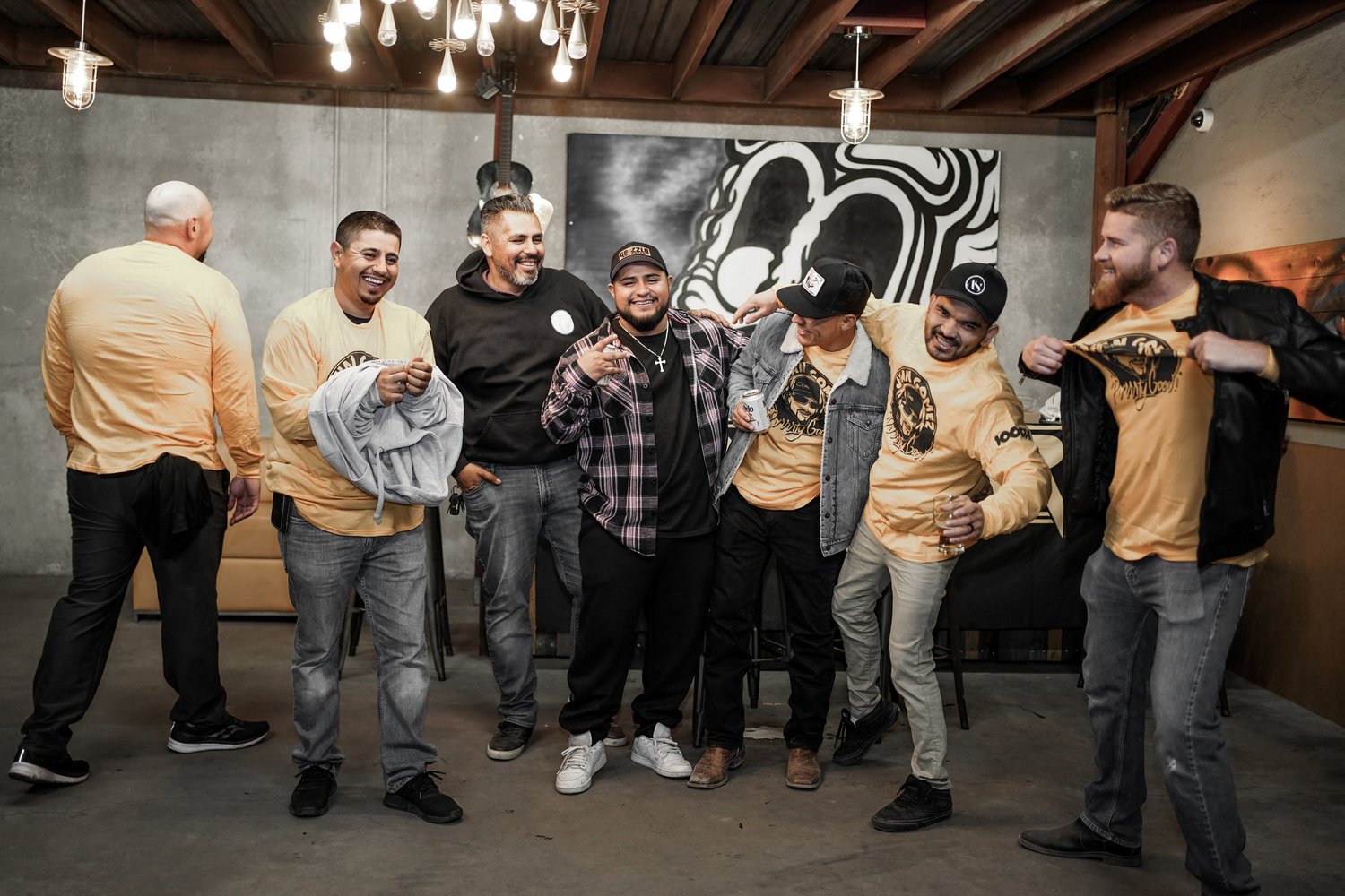 Group of eight men standing together indoors, smiling, and posing for a photo, with exposed wooden ceiling and a large black-and-white abstract art piece on the wall behind them.