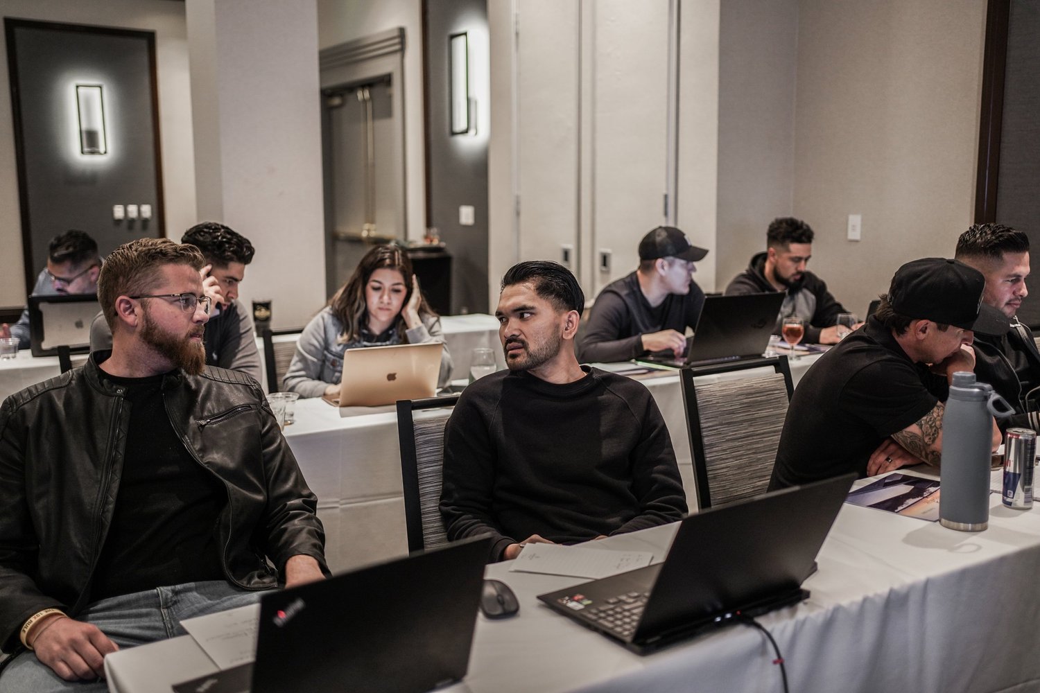 Group of young professionals sitting at a conference table with laptops, engaged in conversation during a meeting or workshop in a modern conference room.