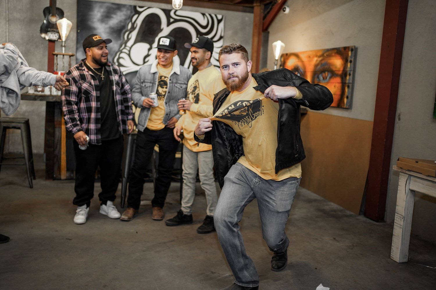 A man with a beard is dancing and pulling open his shirt to show a logo on his T-shirt while a group of four men watch and smile in a party or casual gathering setting indoors.