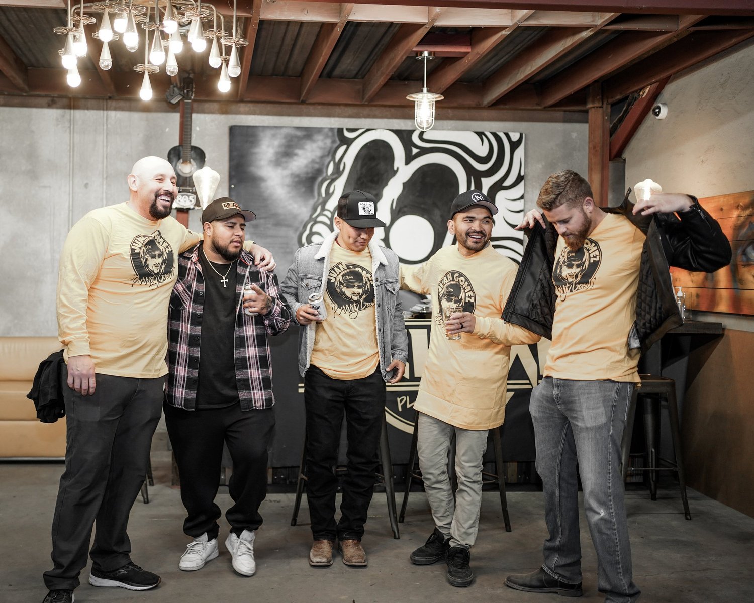 Group of six men standing together indoors, wearing matching yellow T-shirts with a logo, some holding drinks, smiling and interacting with each other.