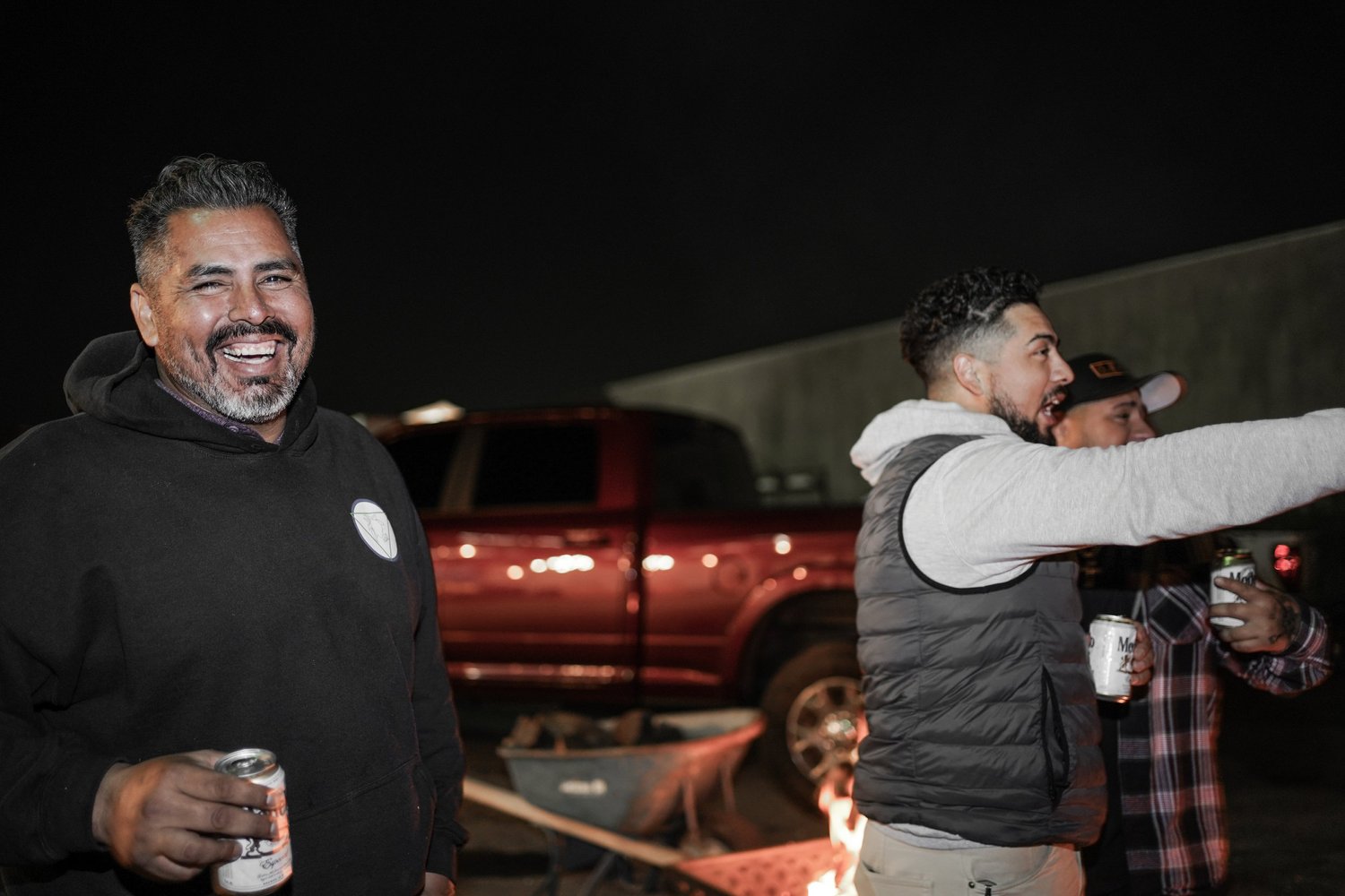 Group of men enjoying a night outdoors, holding drinks, with a red truck and a fire pit in the background.
