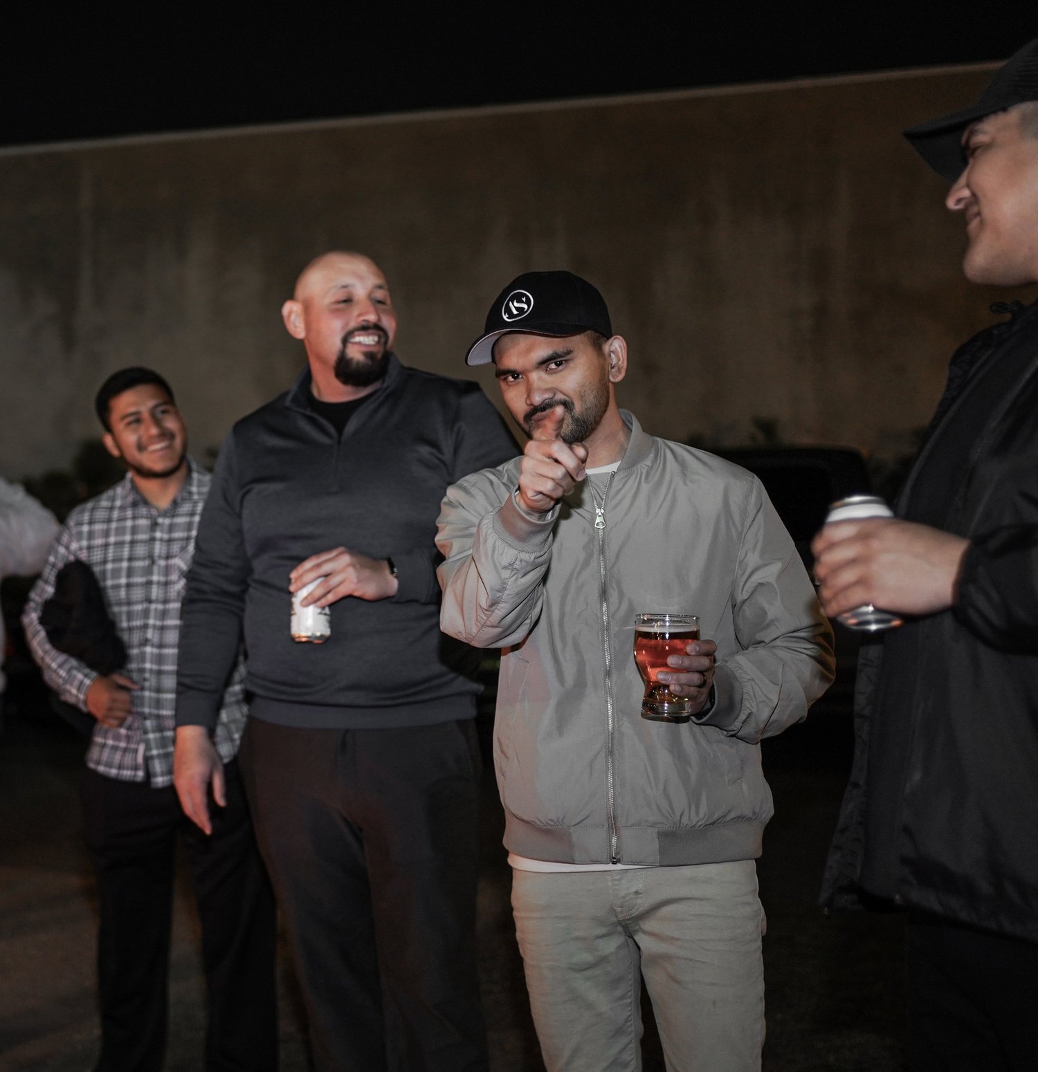 Group of four men socializing outdoors at night, two of them holding drinks, one smiling at the camera, with a dark sky and a large structure or wall in the background.