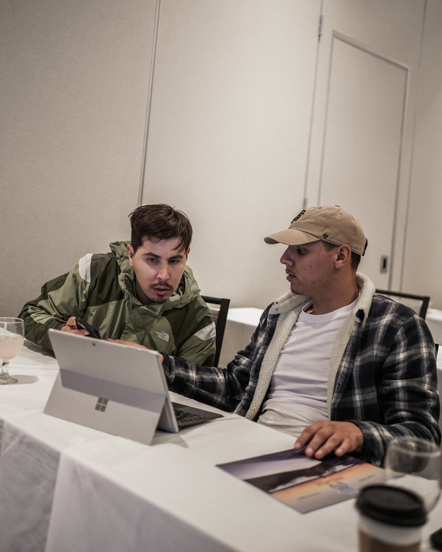 Two young men sitting at a table looking at a tablet, in a room with plain walls. One is wearing a green jacket and holding a phone, the other is wearing a plaid shirt and a tan cap.