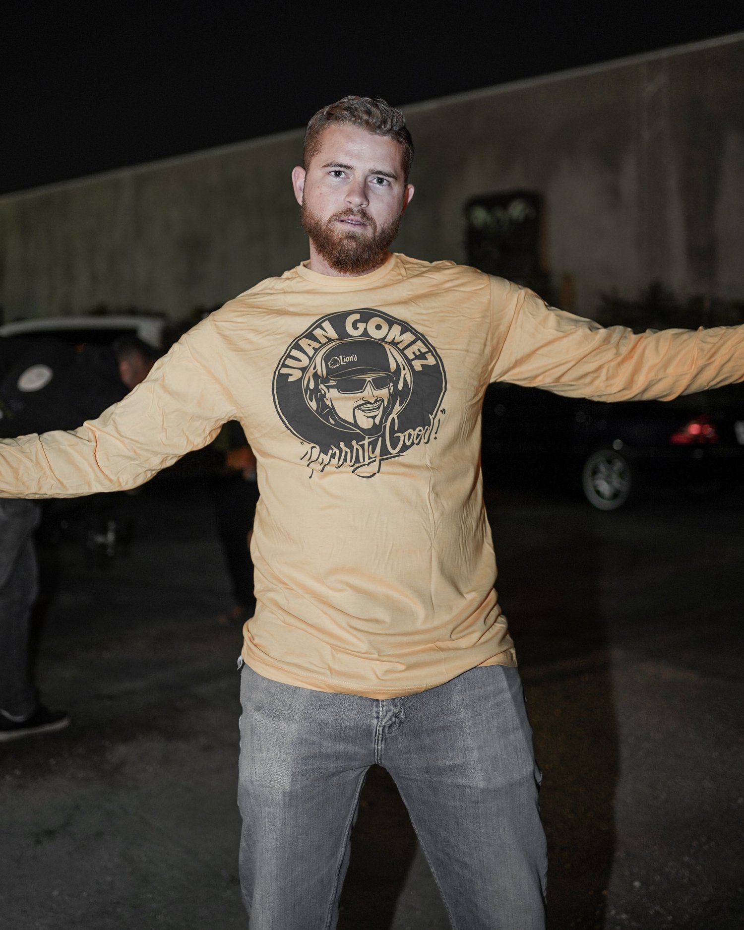 Man with light brown hair and beard wearing a beige shirt with a graphic and text, standing outdoors at night with arms outstretched.
