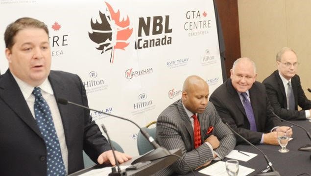 Man speaking at a podium during a press conference with three men sitting at a table behind him. The backdrop displays the NBL Canada logo and various sponsor logos.
