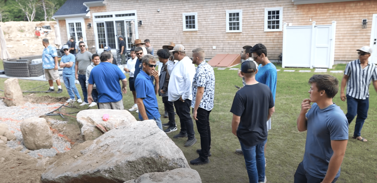 Group of people gathered outdoors in a backyard, observing an archaeological excavation site with large rocks and sand in the foreground.