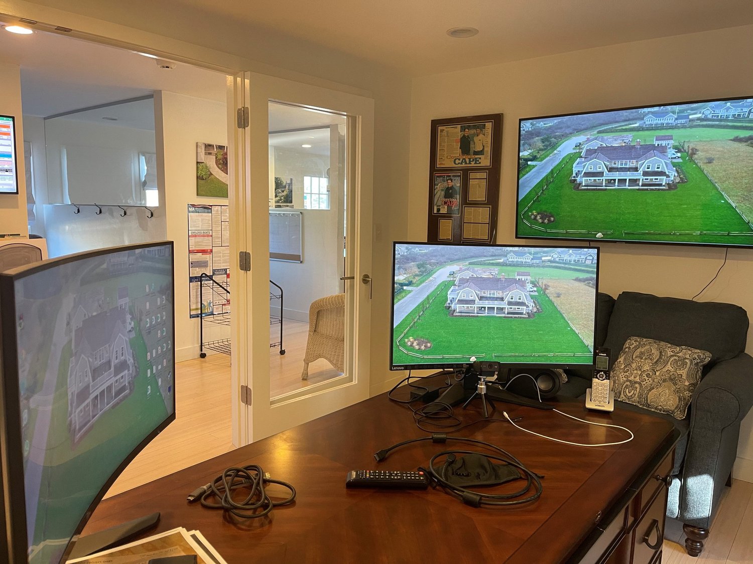 A home office with three screens showing aerial images of a house and yard. The setup includes a curved monitor, a regular monitor, and a TV mounted on the wall. A lamp, remote controls, and cables are on a wooden desk, with a gray armchair nearby.