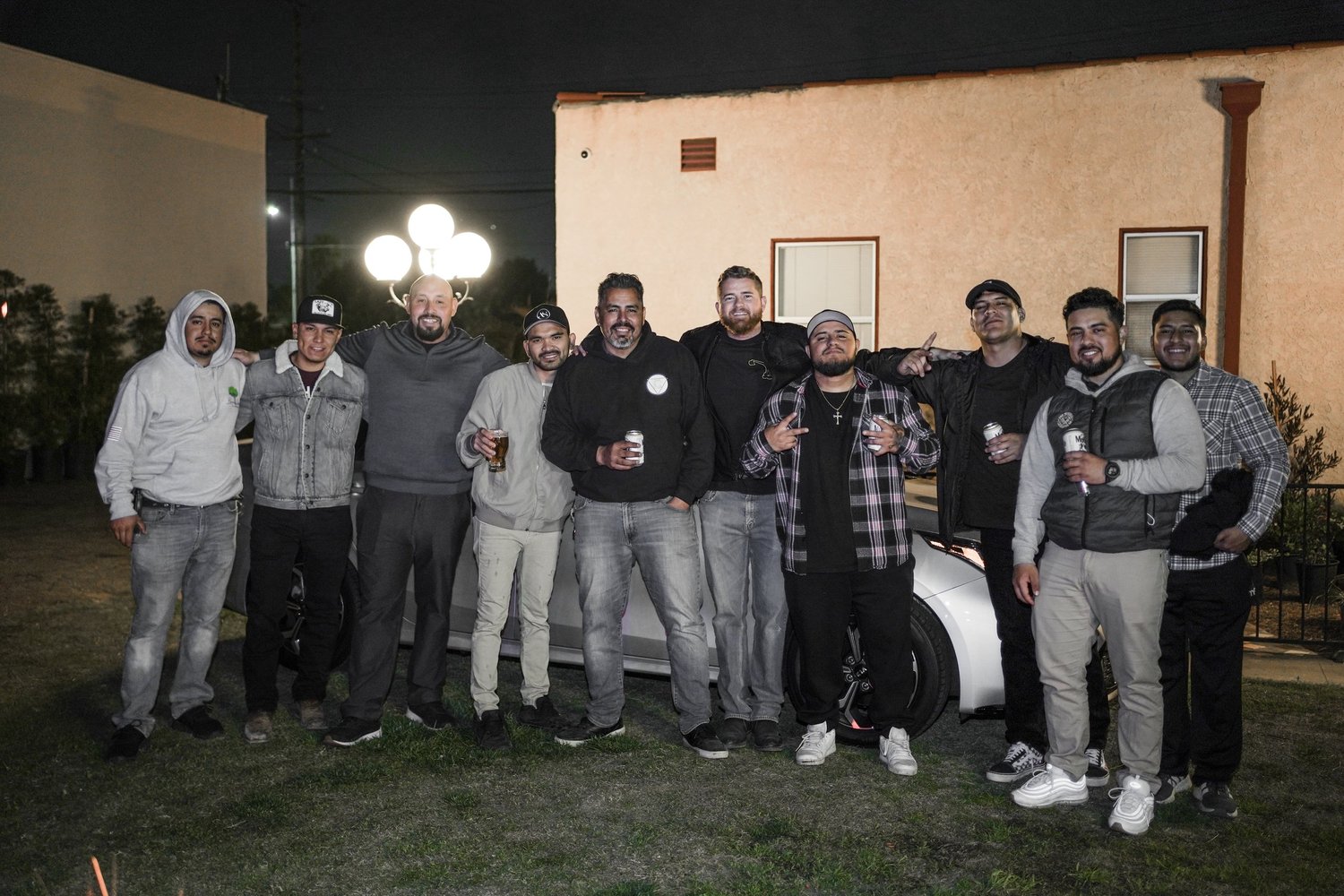 Group of ten men standing outside at night, posing for a photo, some holding drinks, with a white car behind them and a building with a window and a lit lamp post in the background.