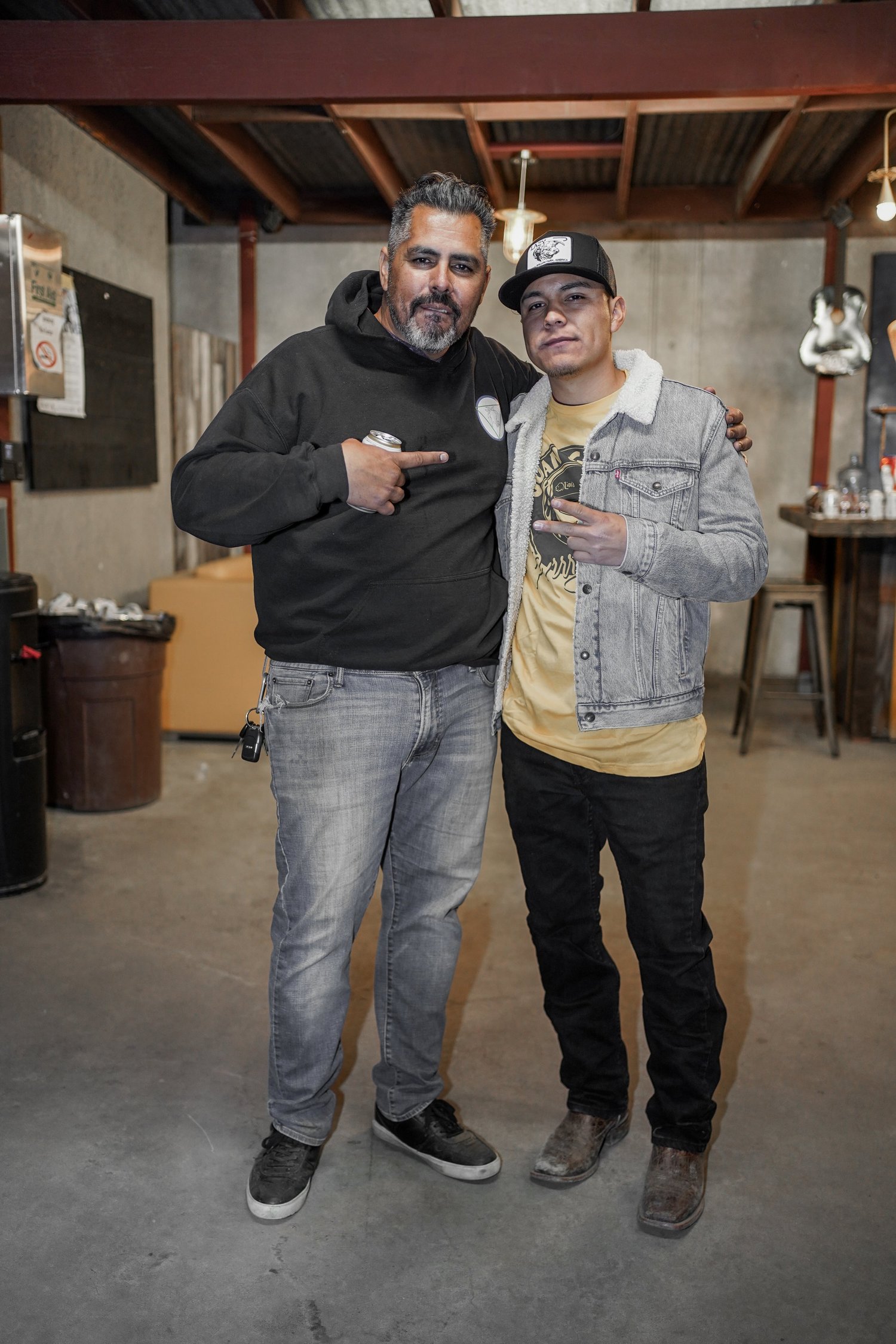 Two men standing together in a rustic indoor setting, smiling and making hand gestures. One man has a beard and is wearing a black hoodie and gray jeans, while the other is younger, wearing a gray jacket, yellow T-shirt, and a baseball cap. The man o